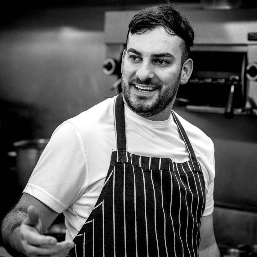 A professional portrait of Chef Scott Hubble wearing a dark chef's jacket, smiling in a kitchen setting.