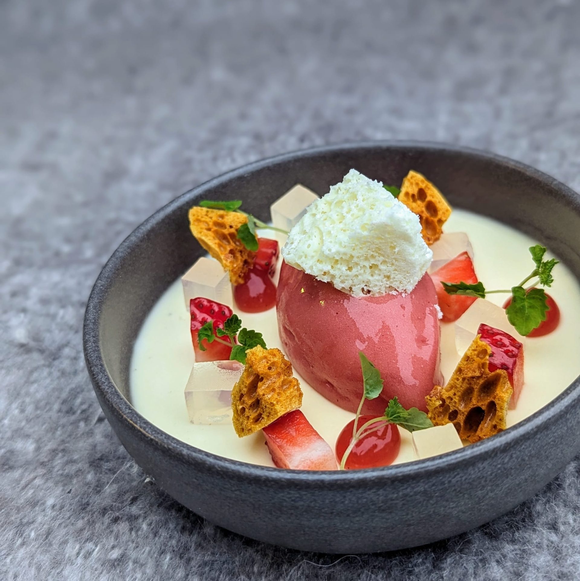 An artistic dessert featuring a fresh strawberry halves, cream, and delicate garnishes on a dark, textured plate.